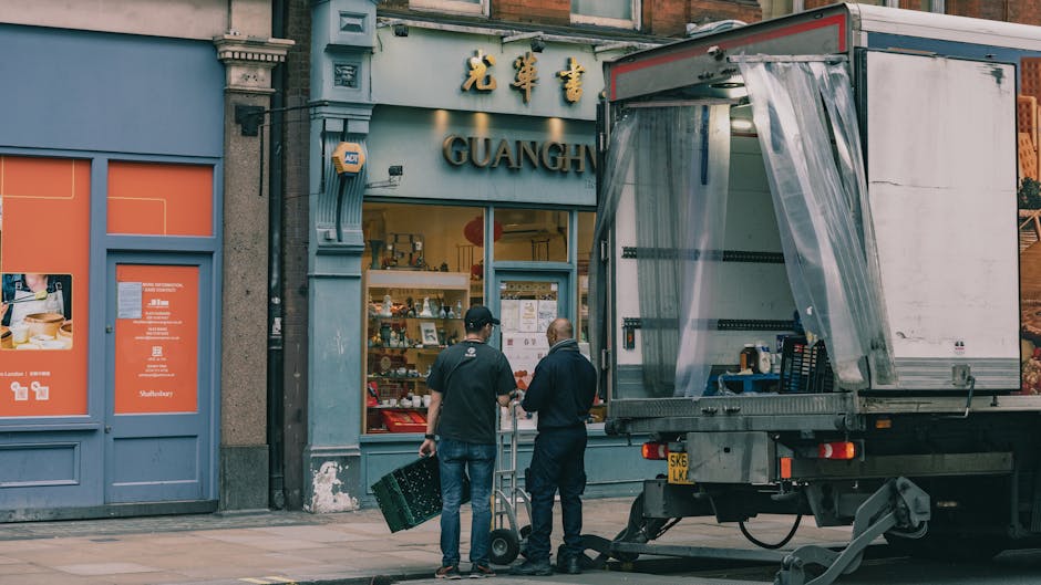 Two removal crew members are outside a commercial storefront with Chinese characters and the word 'GUANGH'. One worker, dressed in dark clothing, is holding a plastic crate, while the other, in a dark