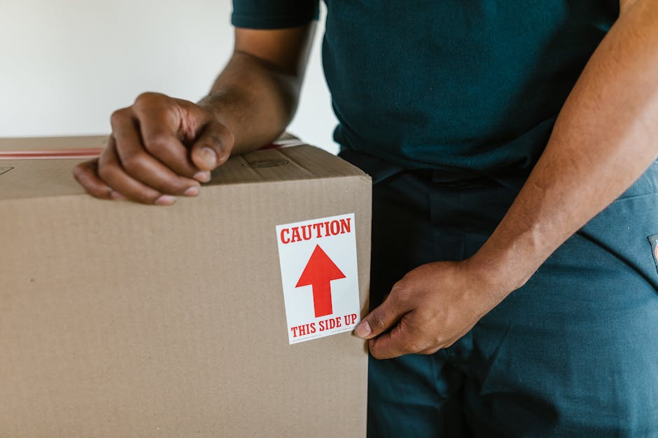 A person wearing a dark short-sleeved shirt and blue trousers is sealing a large cardboard box with a red and white warning label that reads 'CAUTION THIS SIDE UP' with an upward-pointing arrow. The individual is indoors, standing next to a stack of similarly packed boxes, likely preparing for home relocation or furniture transport during a moving process. The background features a plain wall, and the scene is illuminated with natural or neutral lighting, emphasizing the careful handling of packing materials commonly involved in professional removals services like those offered by Man With a Van Clapham. The focus on secure packing and proper handling illustrates key elements of packing and moving logistics aligned with weekend house move timings, supporting effective and safe transport of household items.
