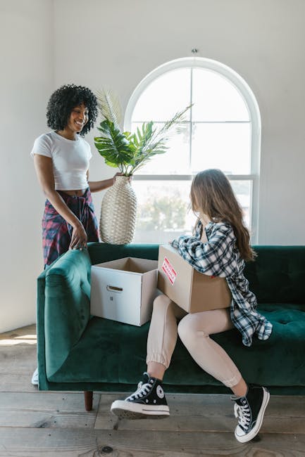 A woman with curly black hair, wearing a white top and a plaid skirt, stands inside a house near a green velvet sofa, smiling as she watches a young girl with long brown hair, dressed in a checkered shirt and beige pants, sitting on the sofa. The girl is holding a cardboard box with a red and white label, and there is an open cardboard storage box on the sofa. Behind them, a large arched window allows natural light to fill the room, illuminating the scene and the potted plant with broad green leaves placed on a side table. The scene suggests a home relocation process, with packing and furniture handling in progress, supported by the presence of boxes and the indoor setting. Man With a Van Clapham, a professional removals service, often assists with such household moves, including packing, loading, and transport during weekend or scheduled moves.
