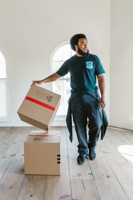 A person wearing a dark short-sleeved shirt and blue trousers is sealing a large cardboard box with a red and white warning label that reads 'CAUTION THIS SIDE UP' with an upward-pointing arrow. The individual is indoors, standing next to a stack of similarly packed boxes, likely preparing for home relocation or furniture transport during a moving process. The background features a plain wall, and the scene is illuminated with natural or neutral lighting, emphasizing the careful handling of packing materials commonly involved in professional removals services like those offered by Man With a Van Clapham. The focus on secure packing and proper handling illustrates key elements of packing and moving logistics aligned with weekend house move timings, supporting effective and safe transport of household items.