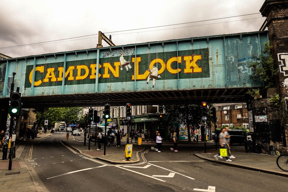 A large, weathered railway viaduct painted in faded blue spans across a busy city street near Clapham Junction station. The structure displays a prominent yellow sign reading 'CAMDEN LOCK' with subtle artwork of climbing figures along its length. Below, there is a pedestrian crossing with people walking and standing on the sidewalks, some wearing casual clothing and carrying bags. Several traffic signals are visible, with green and red lights, and cars and bicycles are moving through the intersection. The street scene includes traffic cones and temporary barriers, indicating ongoing traffic management. The surrounding environment features brick buildings, some of which have graffiti or murals, and trees lining the sidewalk. The lighting is overcast, consistent with cloudy weather typical of urban areas. This scene, with a focus on the moving traffic and pedestrians, reflects typical elements involved in house removals or furniture transport within central London neighborhoods. Man With a Van Clapham provides services that facilitate home relocation and moving logistics in this area, as suggested by the context of busy street movement and urban infrastructure.