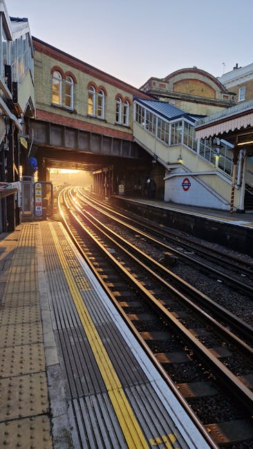The image depicts a railway station platform during late afternoon or early evening, with sunlight illuminating the tracks as they extend toward the horizon. The platform features yellow tactile paving and textured strips along the edge for safety. An overpass level crossing with stairs and glass-fronted structures spans across the railway, connecting the station entrance to the platform area. The surrounding buildings include brick and metal constructions, some with arched windows, indicative of an urban environment near Clapham Junction station. The station appears quiet and clean, with the focus on the railway tracks, station infrastructure, and sunlight creating a warm, inviting atmosphere. This scene relates to house removals and moving logistics, highlighting the importance of efficient transport and freight handling infrastructure in relocation services, as provided by Man With a Van Clapham.