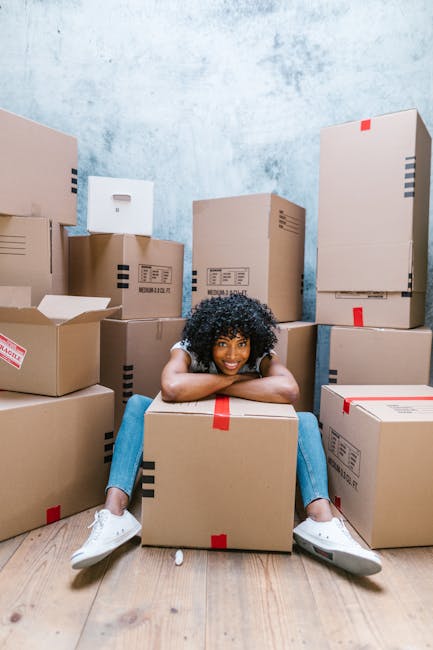 A young woman with curly black hair, wearing a white t-shirt, blue jeans, and white sneakers, is sitting on a light wooden floor surrounded by numerous cardboard boxes of various sizes, some sealed with red and black packing tape and others partially open revealing packing materials. She is smiling at the camera with her arms resting on a large cardboard box in front of her. The boxes are stacked against a textured light blue wall, and some are placed directly on the floor while others are arranged in a semi-organised manner, indicating a home relocation or packing process. The scene suggests an indoor environment, possibly a living room or storage area, with natural or diffused lighting. This image reflects the preparatory stage of furniture transport and packing for a move, in line with the services provided by Man With a Van Clapham as featured on the SW4 removals pricing guide page.