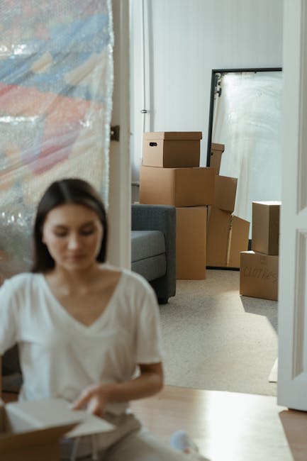Inside a home during a house removal process, a young woman with dark hair and a white t-shirt is seated on the floor, opening a cardboard box. Behind her, there are several packed cardboard boxes of various sizes stacked near a grey sofa, ready for transportation. A large mirror or window leaning against the wall is visible in the background, along with a door to another room. To the left, part of a plastic-wrapped piece of furniture or large item is partially visible. The scene is lit with natural daylight, highlighting the packing and moving activities associated with a home relocation. The setting is a typical living room prepared for furniture transport and interior packing, connecting to the house removals and moving services offered by Man With a Van Clapham, as indicated on the webpage.