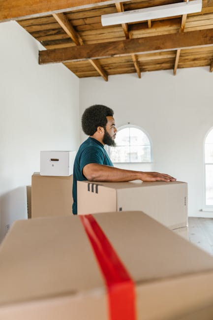 A young woman with curly black hair, wearing a white t-shirt, blue jeans, and white sneakers, is sitting on a light wooden floor surrounded by numerous cardboard boxes of various sizes, some sealed with red and black packing tape and others partially open revealing packing materials. She is smiling at the camera with her arms resting on a large cardboard box in front of her. The boxes are stacked against a textured light blue wall, and some are placed directly on the floor while others are arranged in a semi-organised manner, indicating a home relocation or packing process. The scene suggests an indoor environment, possibly a living room or storage area, with natural or diffused lighting. This image reflects the preparatory stage of furniture transport and packing for a move, in line with the services provided by Man With a Van Clapham as featured on the SW4 removals pricing guide page.